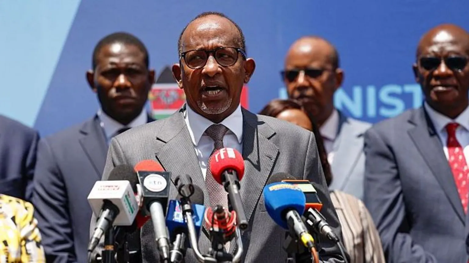 AFP via Getty Images Kenyan Cabinet Secretary of Health Aden Duale in a grey suit, speaks at a podium with microphones, with several people standing behind him