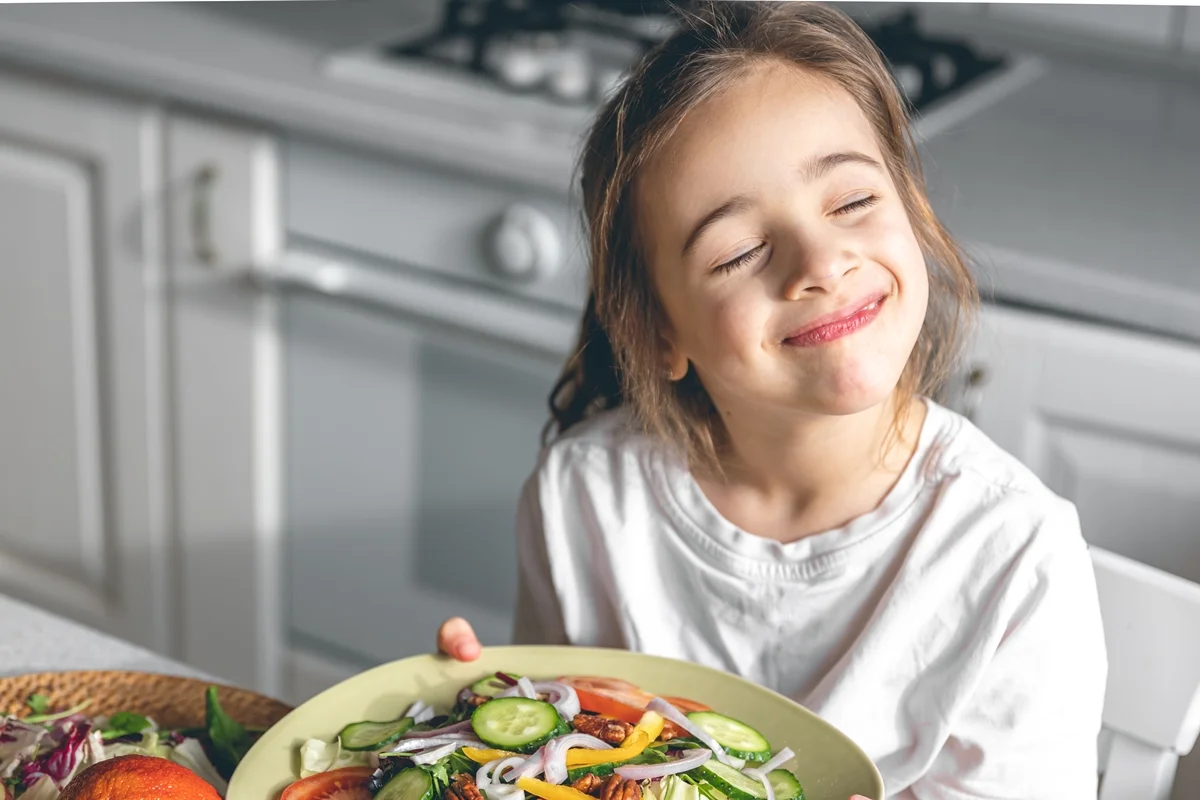 Criança segurando um prato de salada de legumes frescos enquanto está sentada na cozinha; conceito de alimentação saudável. Metrópoles