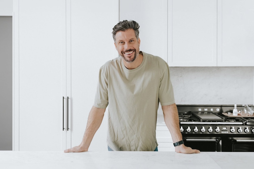 Man in beige t-shirt leaning on kitchen bench with wide smile