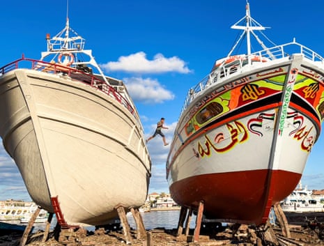 A teenage fisher jumps between two huge wooden boats, one off-white, the other colourfully painted